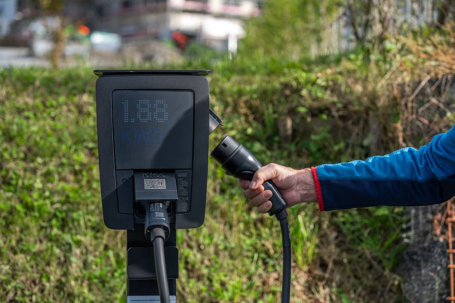 Borne de recharge noire à écran fixée sur un poteau, tenue par une main qui branche un connecteur, devant une haie