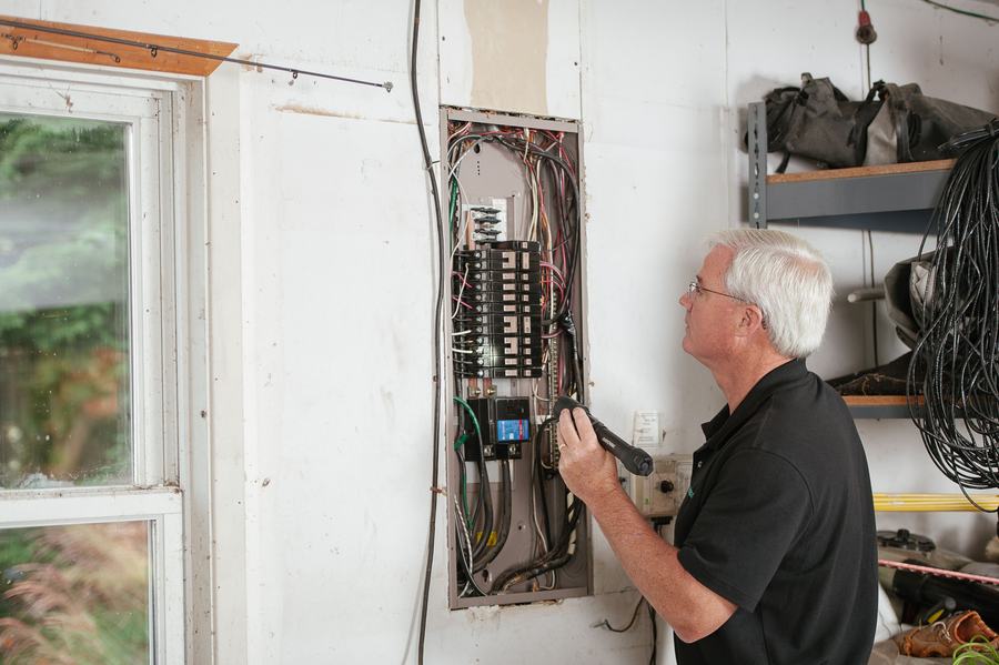 Électricien âgé examinant un tableau électrique encastré dans un mur blanc, près d'une fenêtre